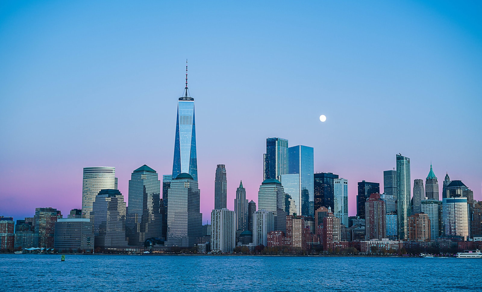 A city skyline at dusk with a full moon rising, the sky a wash of mauve and pink with almost no stars visible.