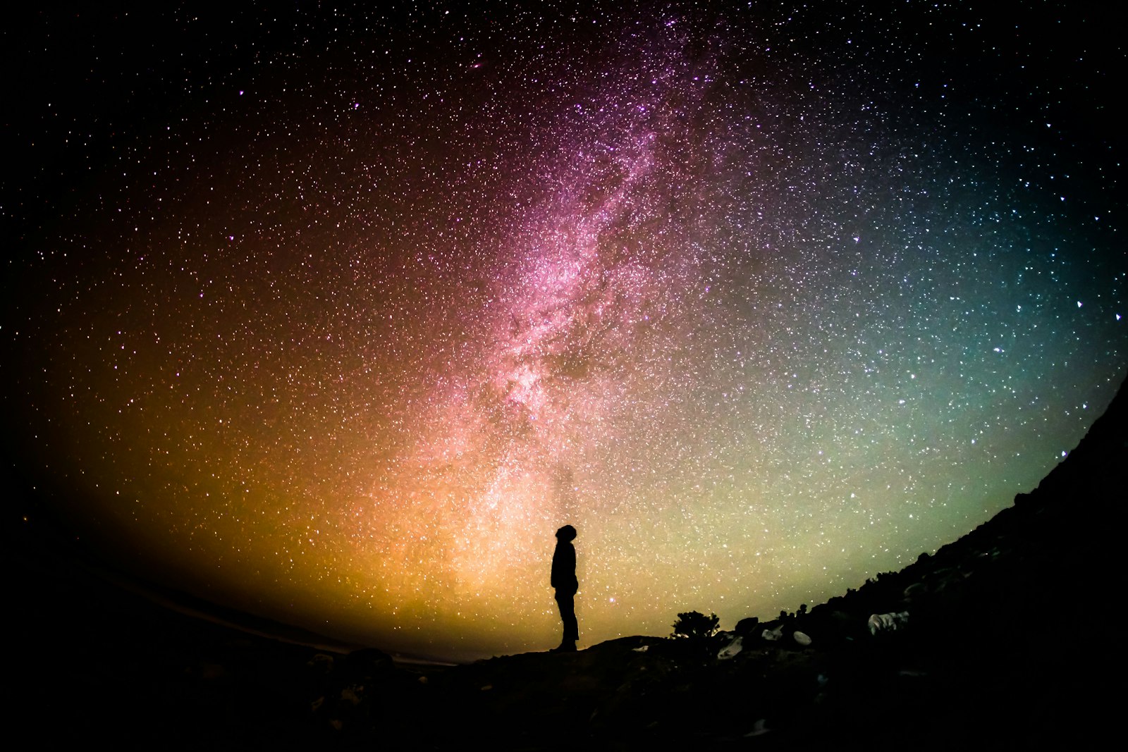 A lone person stands under a vivid, multi-coloured Milky Way that fills the entire sky from horizon to horizon.