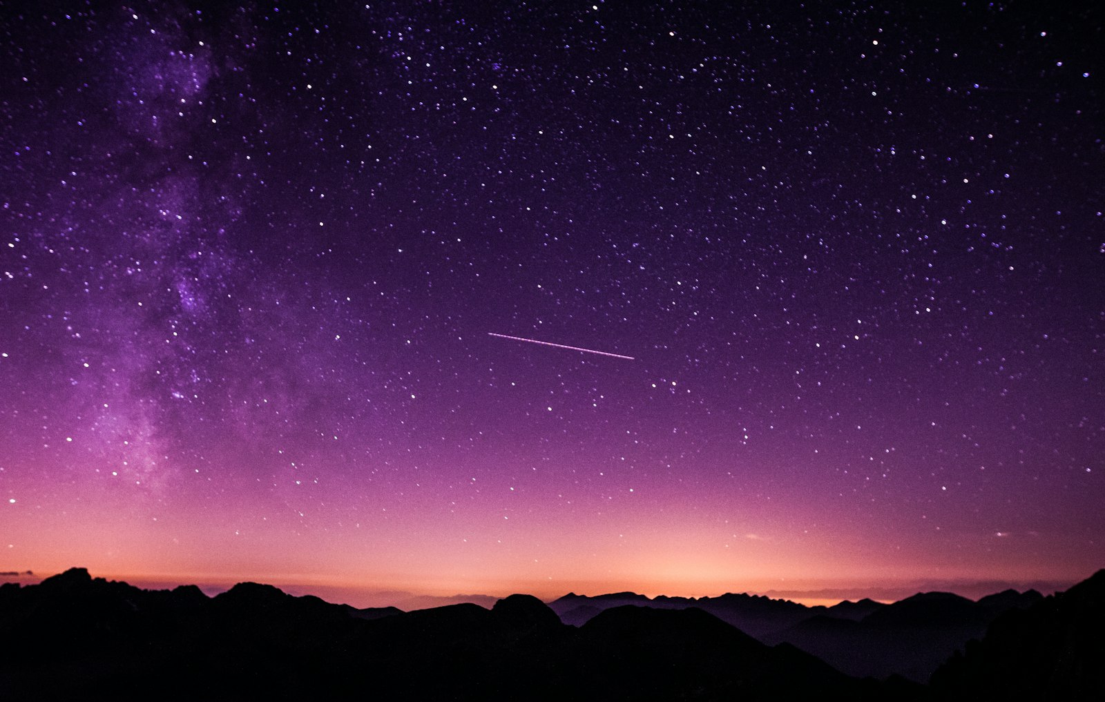 The Milky Way arcing over a mountain horizon at dusk, the sky shading from deep violet to warm amber near the ground.
