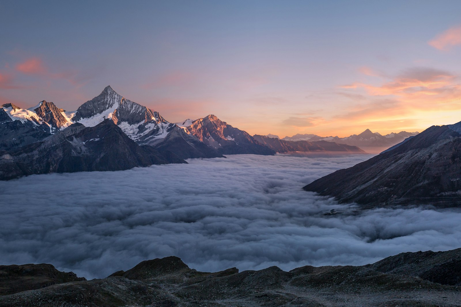 A warm sunset glowing over mountain peaks with a sea of cloud below, the sky fading from orange to dusky blue.