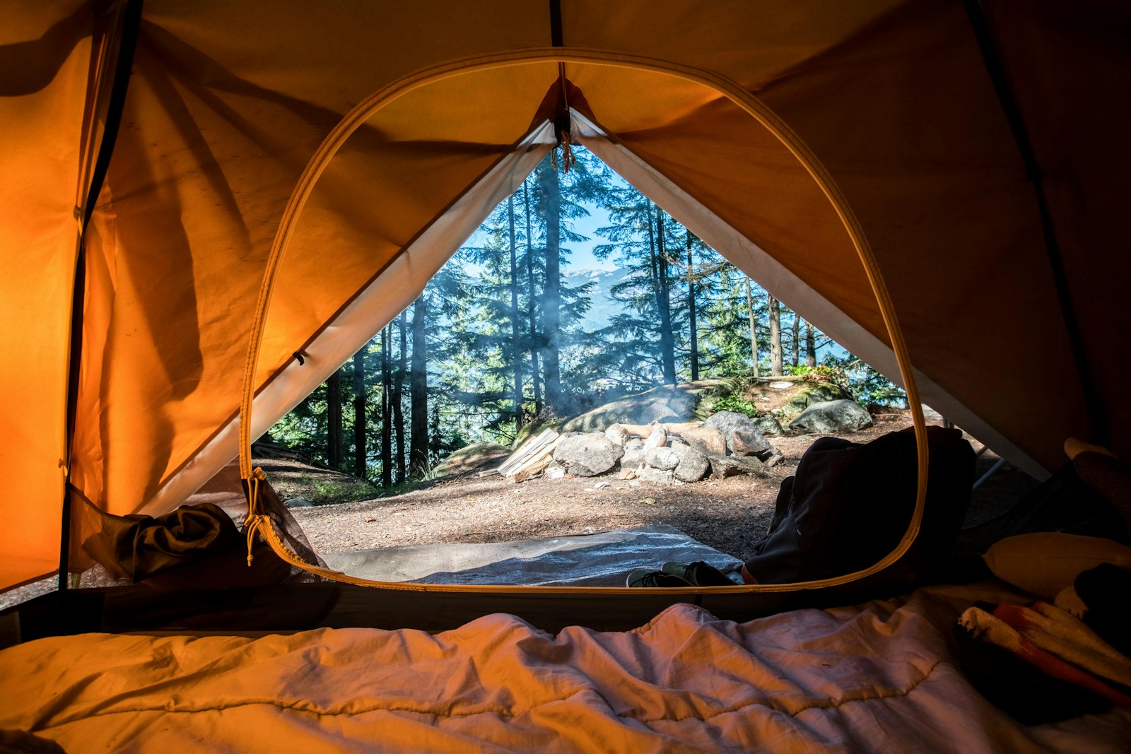 The warm glow inside an orange tent at a forest campsite, looking out toward pines and a smouldering campfire.
